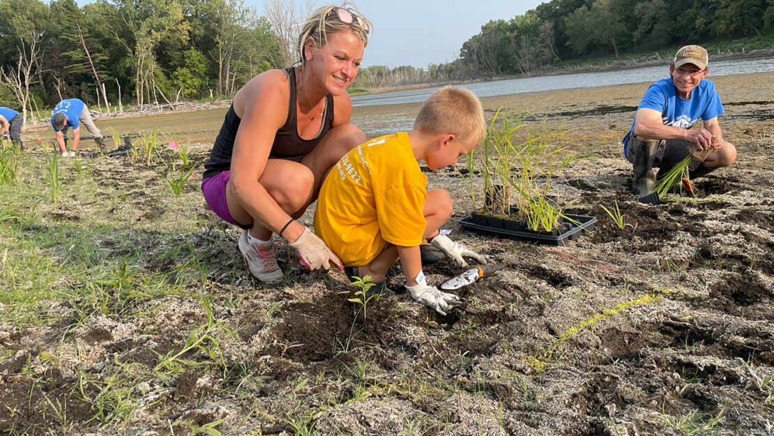 An adult helps a child plant along the shoreline of a wetland.