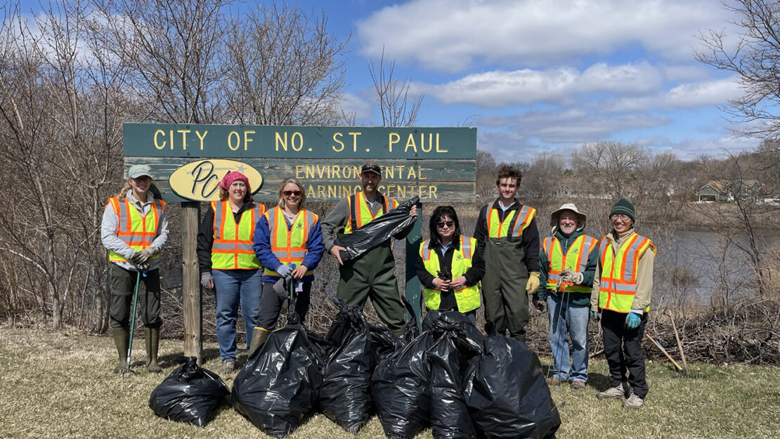 A group of 8 people stand near PCU Pond in North St. Paul with a pile of collected garbage.