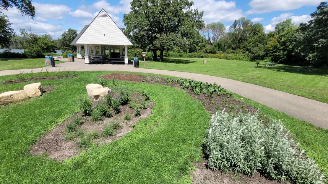 A small white pavilion with a triangular roof sits among lush greenery and a winding path. Vibrant gardens and scattered trees create a tranquil park scene.