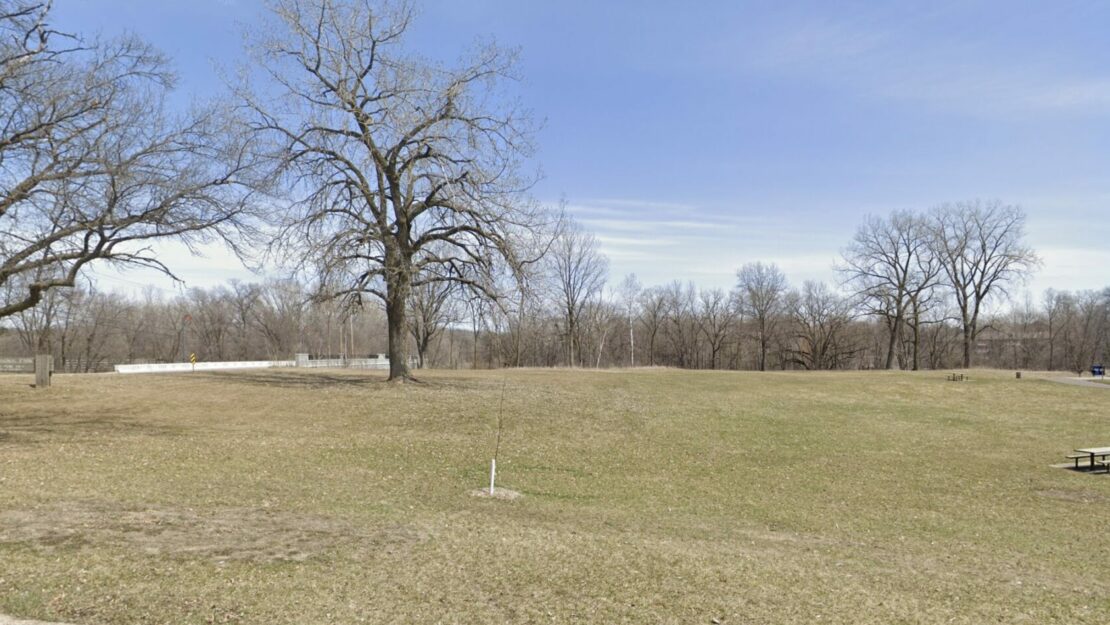 Open field with sparse brown grass and large bare trees under a clear blue sky. A picnic table sits isolated on the right