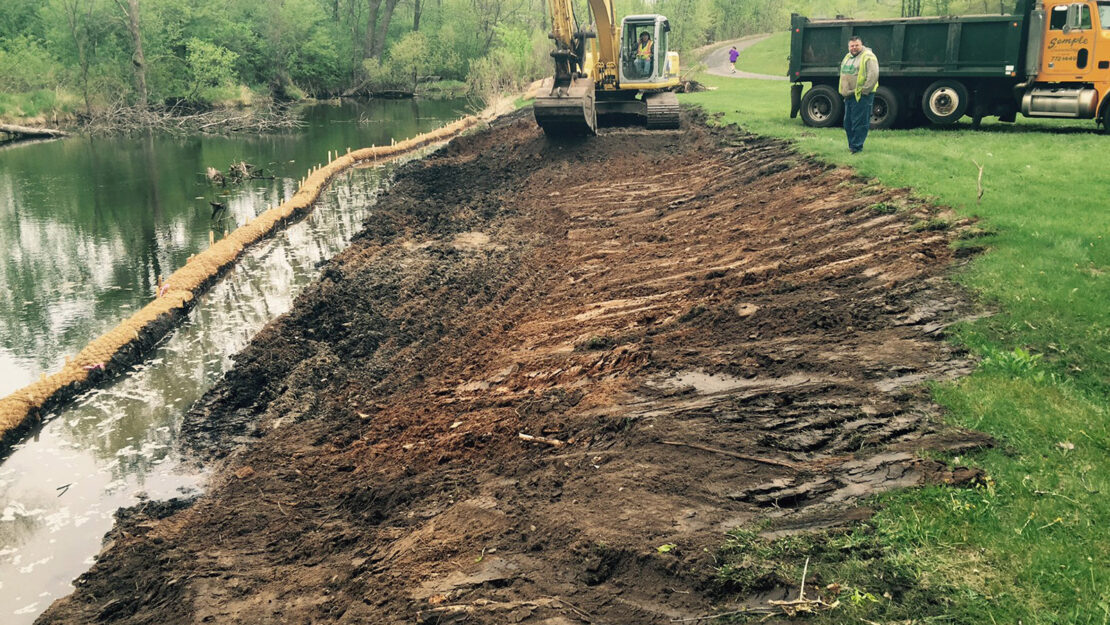 Excavator working on a muddy riverbank, with a silt barrier in the water. A person stands near a yellow truck, surrounded by lush green trees.