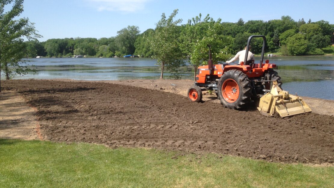 A person on a red tractor tills soil by a lakeside. The scene is calm, with green trees and a clear blue sky.